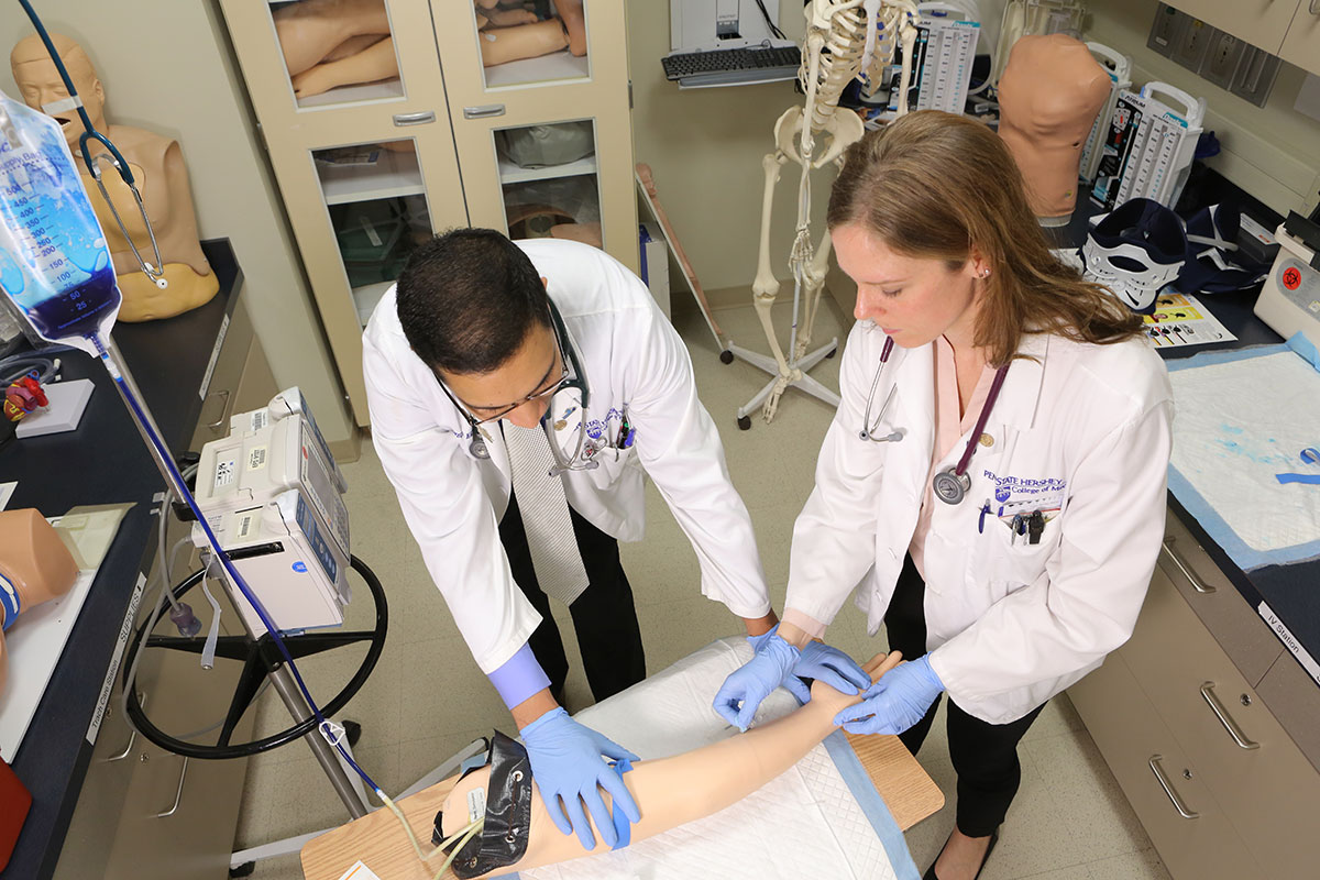 Students Zainul Hasanali and Lauren Kaminsky in the Penn State College of Medicine MD/PhD Program practice a simulation exercise in the Penn State College of Medicine Simulation Center in April 2017. The students are seen wearing short white lab coats and blue gloves, working with a model arm, which is in the lower center of the photo with an IV bag to the left.