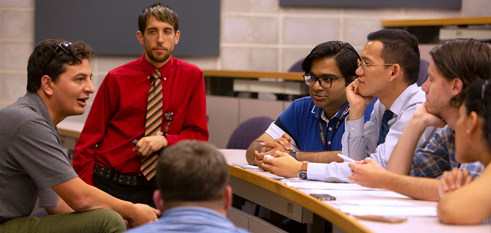 A group of medical students are seen in a lecture room, talking together. Dr. Joshua Davis of Penn State College of Medicine is seen in the back, looking over the group.
