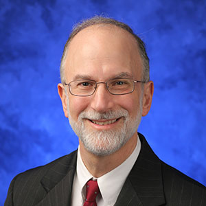 Aron Lukacher, MD, PhD, is Chair of the Department of Microbiology and Immunology at Penn State College of Medicine. He is pictured in a dark suit jacket and red tie against a blue background.