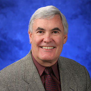 James Broach, PhD, is Chair of the Department of Biochemistry and Molecular Biology at Penn State College of Medicine. He is pictured in a tan suit jacket, maroon shirt and dark tie against a blue background.