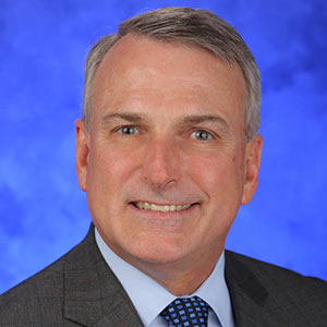 Kent Vrana, PhD, is Chair of the Department of Pharmacology at Penn State College of Medicine. He is pictured in a dark suit jacket, light blue shirt and blue patterned tie against a blue background.