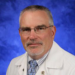 Ronald Wilson, VMD, MS, DACLAM, is Chair of the Department of Comparative Medicine at Penn State College of Medicine. He is pictured in a white lab coat, light blue shirt and light blue patterned tie against a blue background.