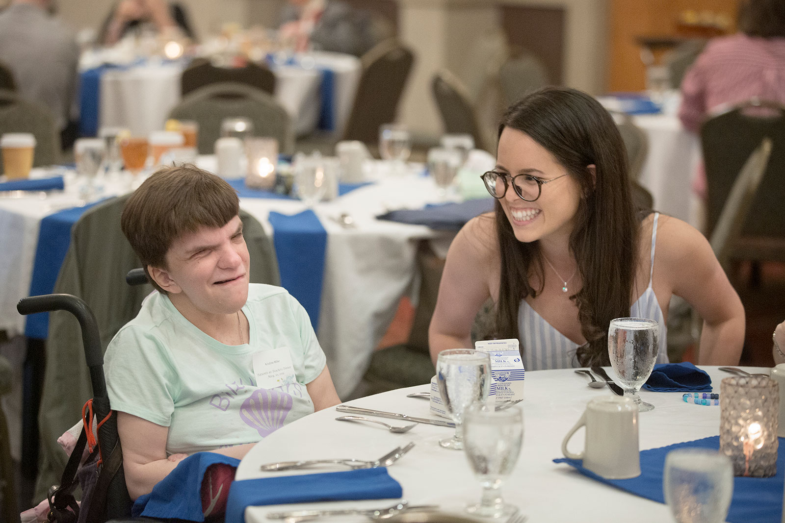 A young woman is seen talking happily with a woman in a wheelchair sitting at a table with her.