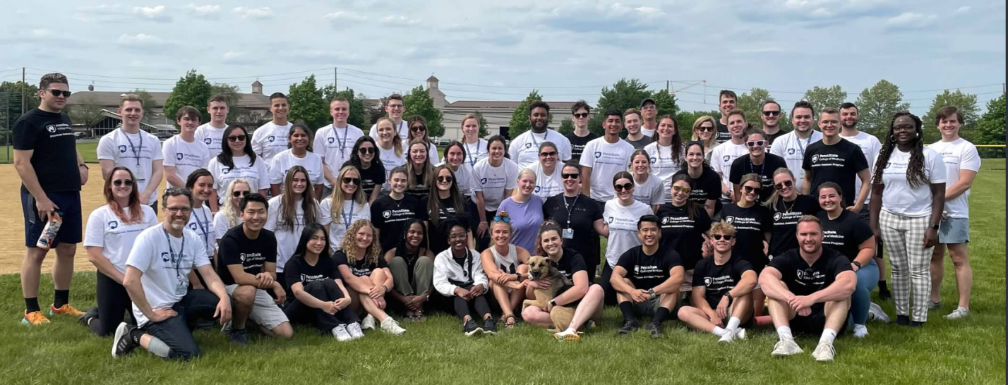 A large group photo of more than 50 people in white and black Penn State College of Medicine shirts on the front lawn of the college in Hershey.