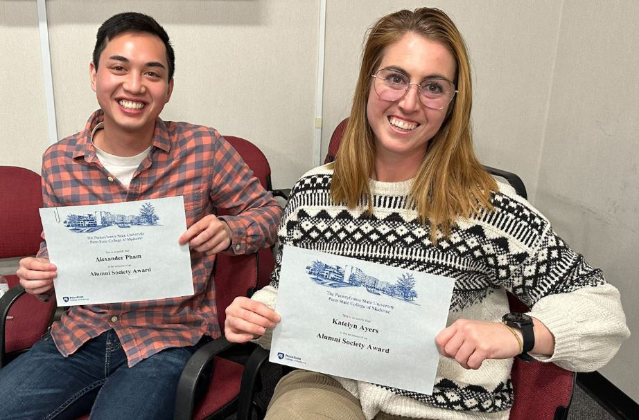 Graduate students Alexander Pham and Katelyn Ayers sit in chairs and hold up their awards