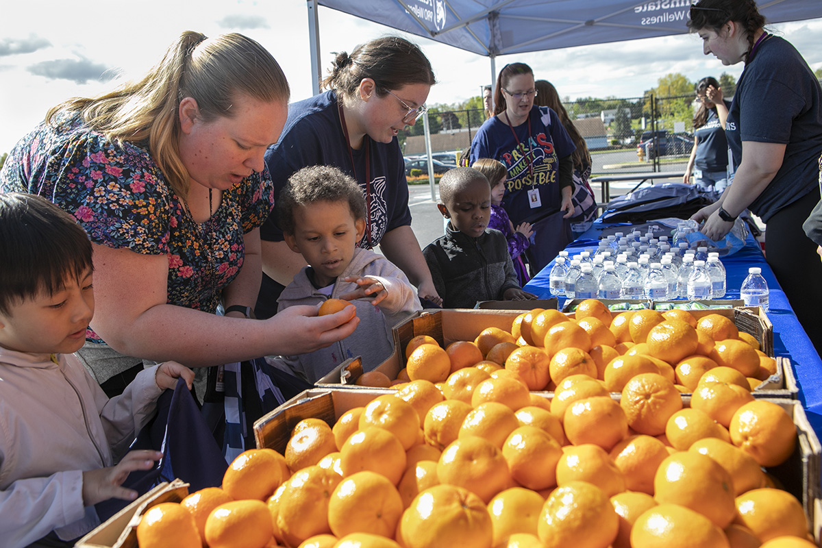 A woman helps several children get fruit from a box on a table under a tent; in the background are water bottles.