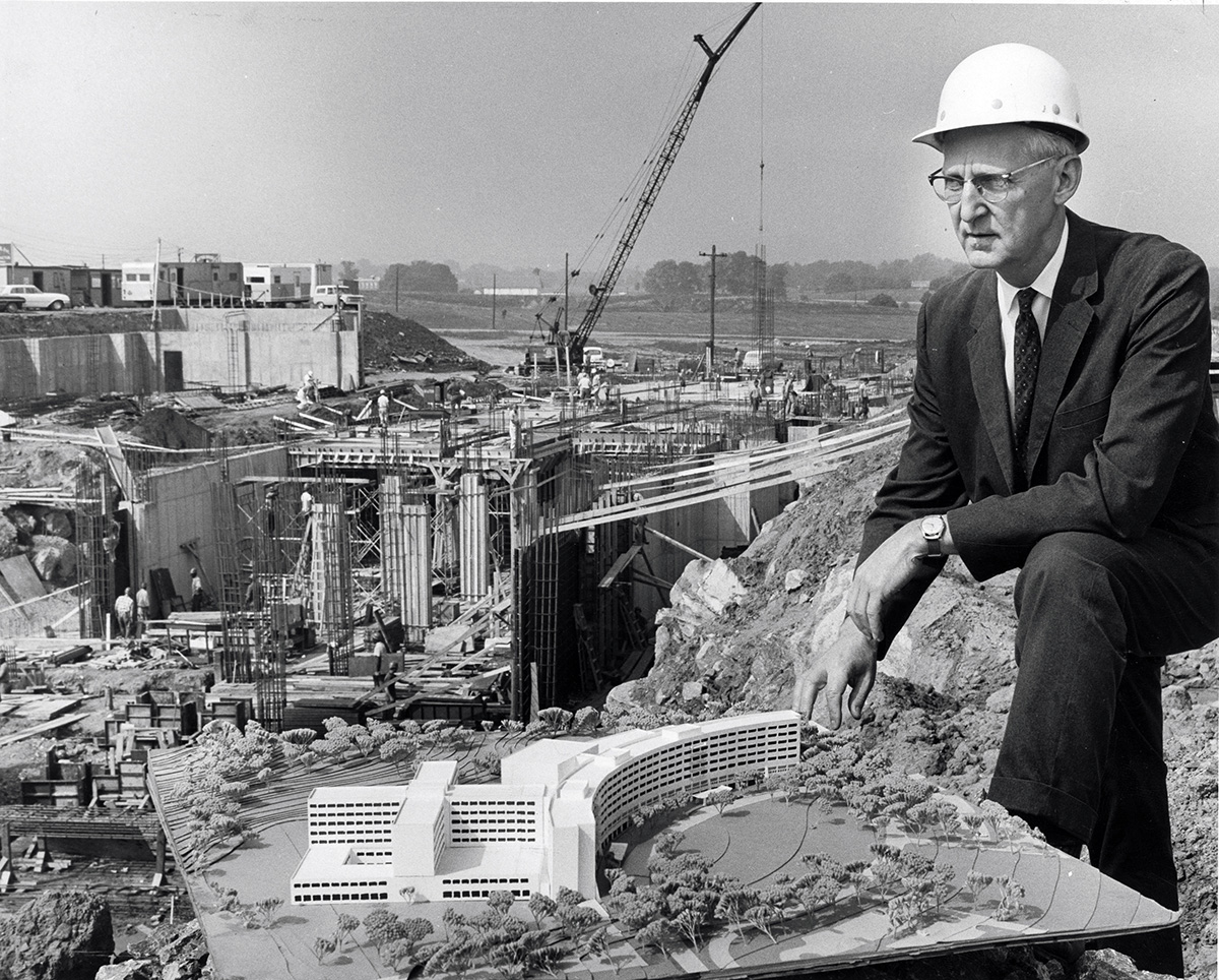 George T. Harrell, MD, in a hard hat and suit, sits with a model of the planned College of Medicine; construction is happening in the background.