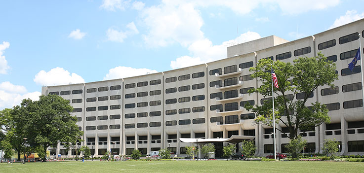 The Penn State College of Medicine Crescent is seen at 500 University Drive, Hershey, PA. The building is framed by grass below and a bright blue sky with white clouds above. Flagpoles with the American and Pennsylvania flags are visible toward the right.