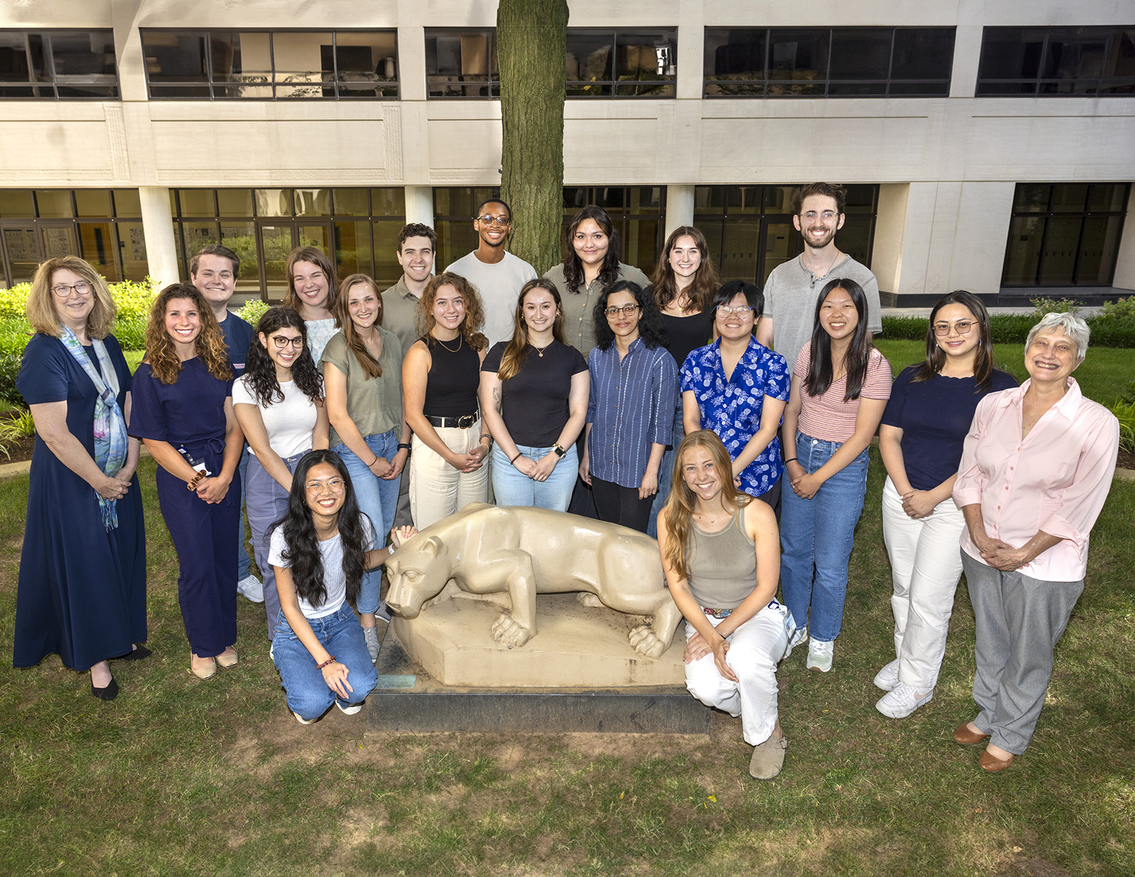 A group of 20 people pose in the College of Medicine courtyard around the Nittany Lion statue; two are kneeling or seated at the sides of the statue and the rest are standing behind and to the side.