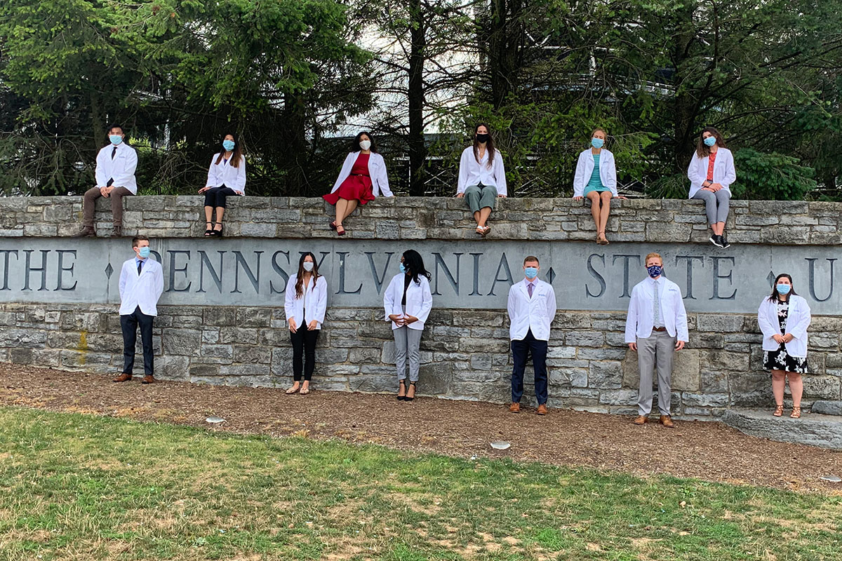 A group of 12 students are pictured, socially distanced and wearing masks, sitting on and standing in front of a wall in which the words The Pennsylvania State University are engraved.