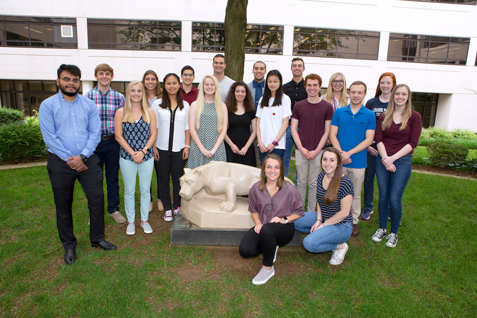 Student participants in the Summer Undergraduate Research Internship Program, or SURIP, at Penn State College of Medicine are seen in 2018. The large group of students is pictured standing in a courtyard at the College of Medicine, with trees, grass and a white building in the background and a statute of the Penn State Nittany Lion mascot in front of them.