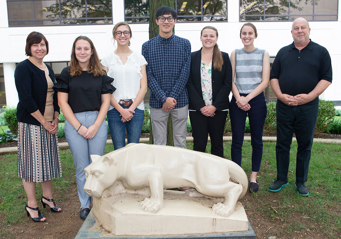 Student participants in the Schreyer Honors College MD/PhD Summer Exposure Program at Penn State College of Medicine are seen in 2018. The group, comprising five students and two faculty members, is pictured standing in a courtyard at the College of Medicine, with trees, grass and a building in the background and a statute of the Penn State Nittany Lion mascot in front of them.