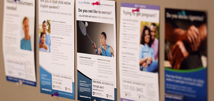 Posters advertising a variety of clinical research trials at Penn State College of Medicine are seen on a College bulletin board in summer 2016. The image shows five posters in a line, with the center one in focus and the others out-of-focus in the background.