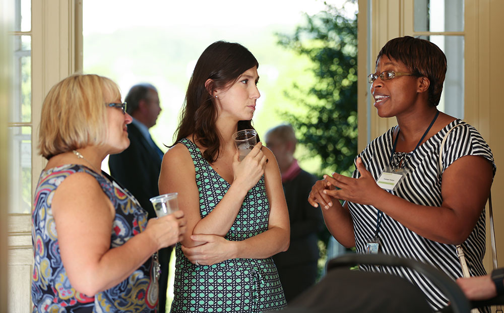 Three women attending a Summer 2016 Innovation Cafe event on female entrepreneurs in medicine are pictured talking with each other. The two women at left are holding plastic cups while looking at the woman at right, who is speaking and gesturing with her hands. Other attendees at the event are pictured out-of-focus in the background in a wide doorway, framed by a tree and the open doors.