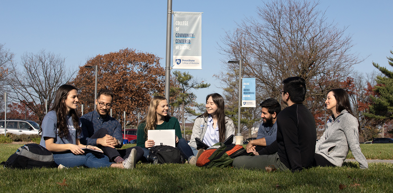 Seven students sit and talk with notebooks and backpacks on the grass outside the College of Medicine; a pole banner says 'College of Community-Centered Medicine'