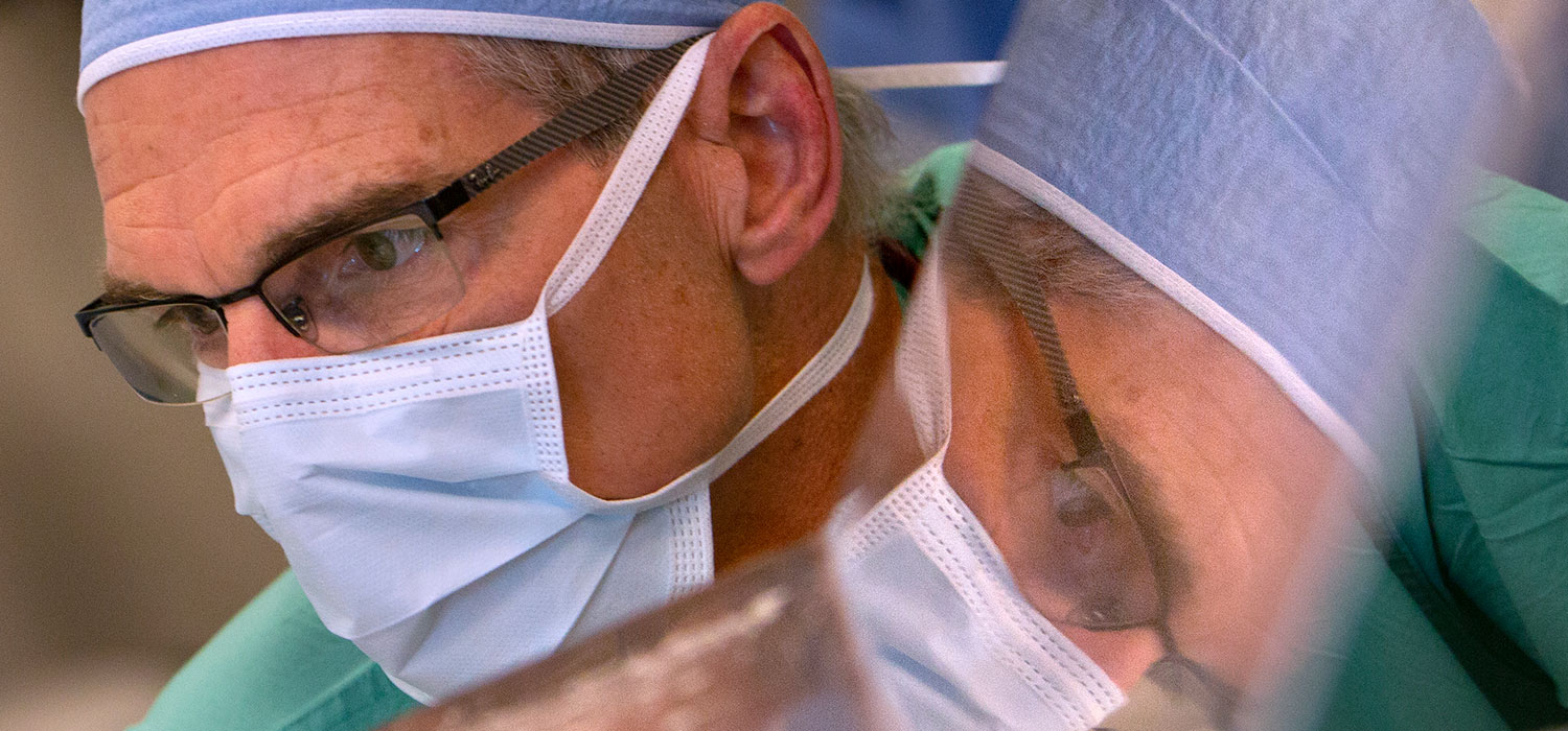 Dr. Berend Mets, Chair of the Department of Anesthesiology, is seen in at work an operating room in May 2018. A close-up view of his face is seen, wearing a surgical hat and mask; his reflection is visible to one side.