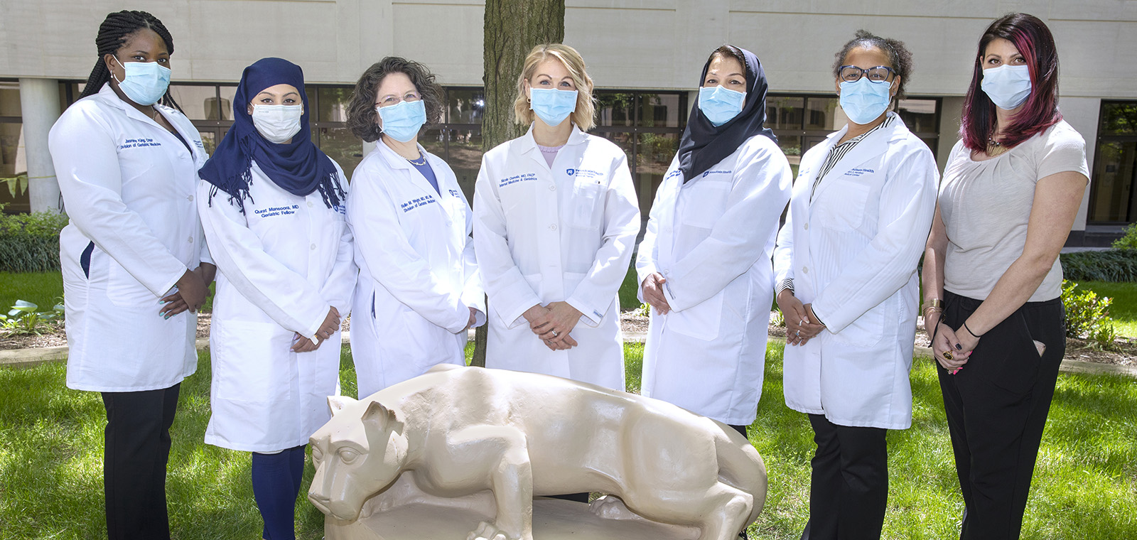 A group of 7 women from the Department of Medicine's Division of Geriatric Medicine stand outdoors, with trees in the background and a lion statue in the foreground, wearing white coats, surgical masks and business professional clothing.
