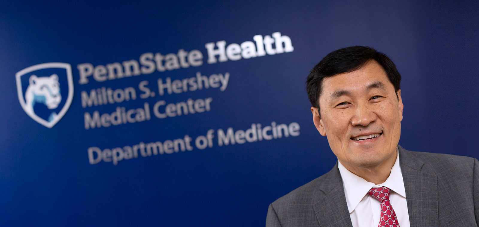 Dr. Thomas Ma, chair of the Department of Medicine at Penn State College of Medicine, is pictured in front of a blue photo backdrop with the department's logo. He is talking animatedly and looks happy.