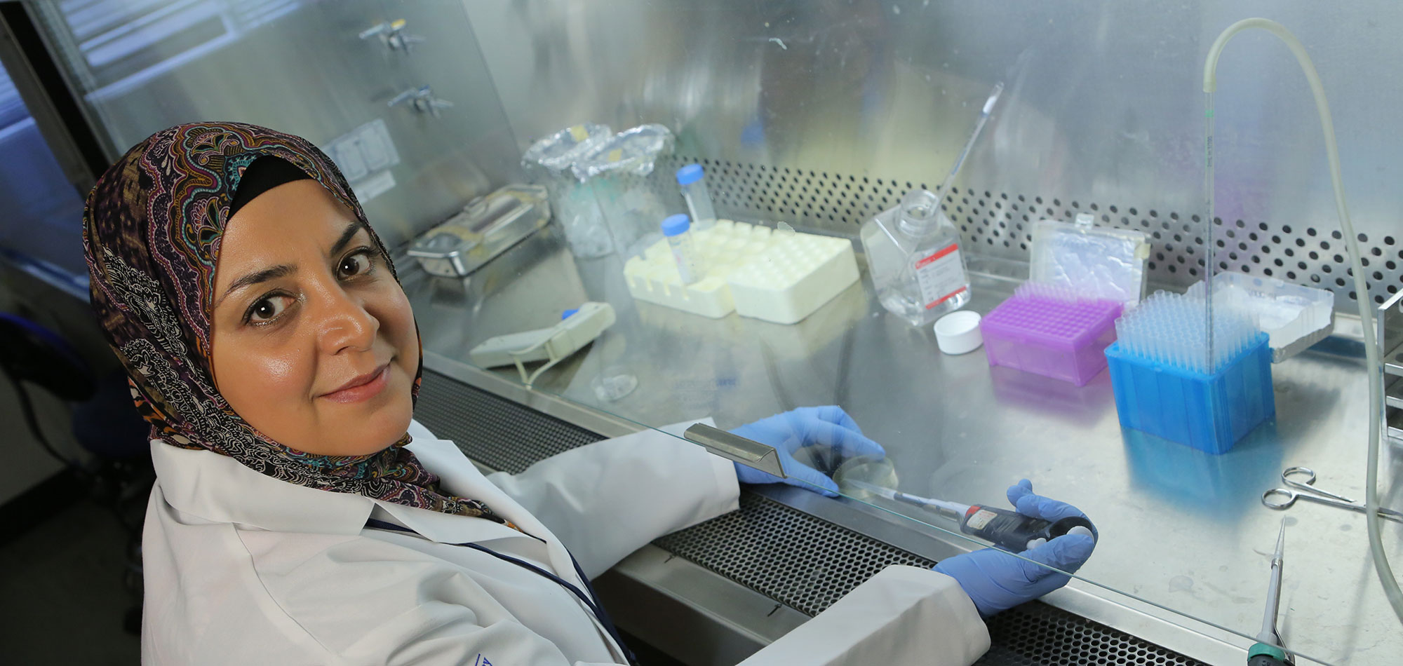 Dr. Fadia Kamal works in a lab in the Department of Orthopaedics and Rehabilitation at Penn State College of Medicine in August 2017. She is holding a pipette in one hand and a small dish in the other and looking toward the camera.