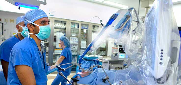 David Goldenberg, MD, FACS, otolaryngology (head and neck) surgeon at Penn State Health Milton S. Hershey Medical Center, is seen in the operating room at the Medical Center in 2015, operating the Flex robot. He is pictured wearing a surgical mask and blue surgical scrubs, with the side view of another surgeon in scrubs seen to the left of the photo.