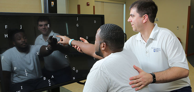An employee of Penn State Physical Medicine and Rehabilitation works with a client at Penn State Rehabilitation Hospital in Hummelstown, PA, in early 2017. The client is seated and pointing at a digital screen with letters on it, and the employee is guiding him.