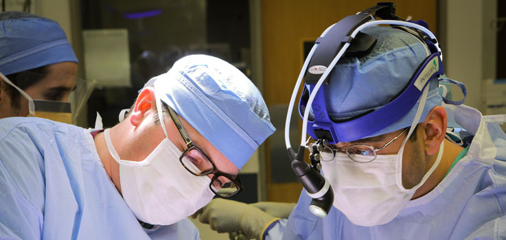 Matthew Taylor, MD, thoracic surgeon and Alok Dash, MD, fellow, thoracic surgeon at Penn State Health Milton S. Hershey Medical Center, is seen in the operating room at the Medical Center in 2016, operating the Flex robot. They are pictured wearing a surgical mask and blue surgical scrubs, with the side view of another surgeon in scrubs seen to the left of the photo.