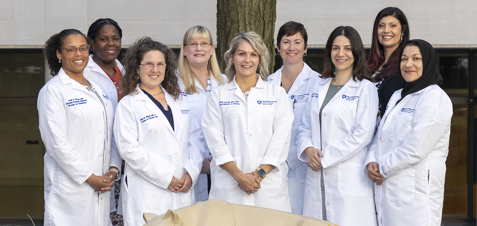 Nine members of the Division of Geriatric Medicine, eight wearing physicians' white coats, stand behind the Nittany Lion statue in the College of Medicine courtyard.