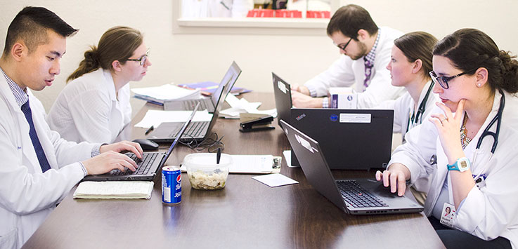 Medical students at Penn State College of Medicine are seen sitting around a table in their white coats, working at laptops, depicting the administrative aspects of the healthcare process.