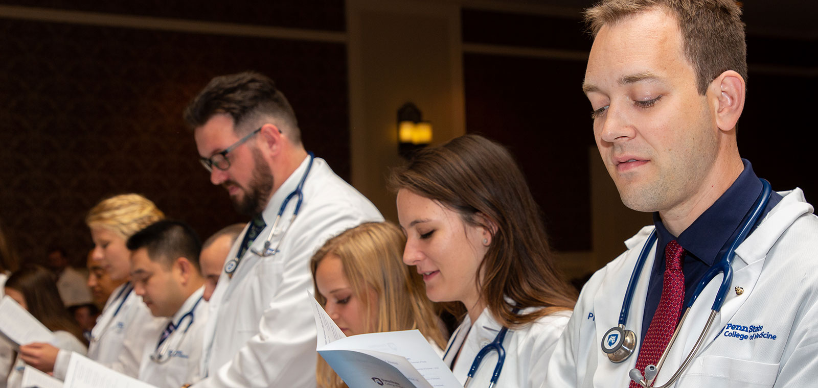 Students in the Physician Assistant Program at Penn State College of Medicine take part in the White Coat Ceremony in 2016, during which the students receive their white coats signifying the start of their training. Three female students are seen in the foreground, reading from the event program, with a male and female student visible in the background.