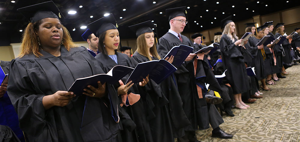 Penn State graduate students from the Department of Public Health Sciences attend the College of Medicine Commencement Ceremony at the Hershey Lodge on May 20, 2018. The students are pictured standing in caps and gowns, holding programs.