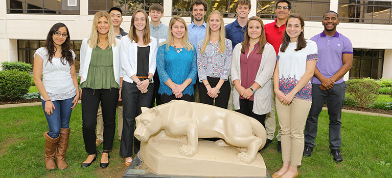 Students from the 2017 SURIP (Summer Undergraduate Research Internship Program) at Penn State College of Medicine are pictured standing near the statue of the Nittany Lion in the College courtyard. An academic building and grass can be seen behind the group of more than 10 students.