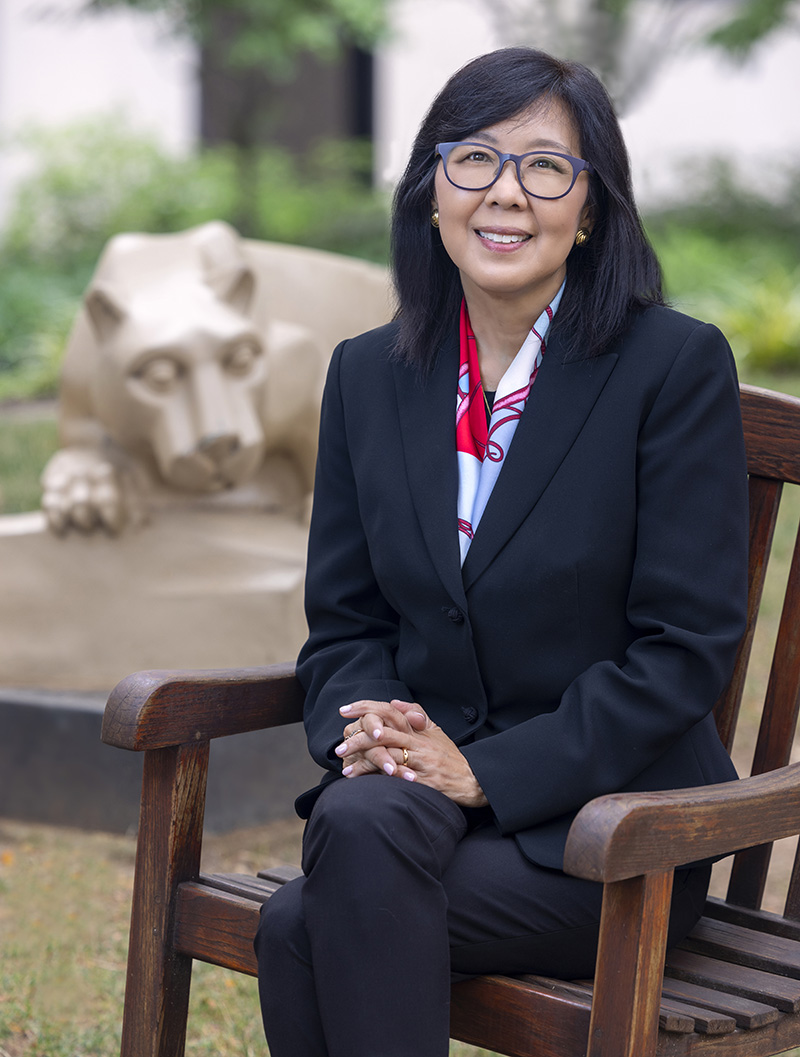 Dr. Karen Kim, dean of Penn State College of Medicine, sites in a wooden chair in a courtyard with the Nittany Lion statue behind her 