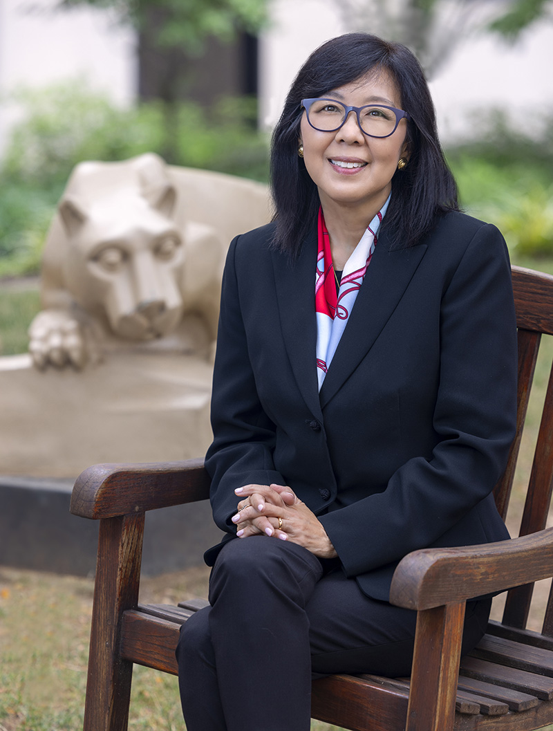 Dr. Karen Kim, dean of Penn State College of Medicine, sites in a wooden chair in a courtyard with the Nittany Lion statue behind her