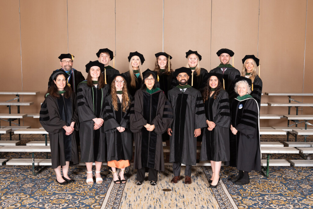 A group of 14 people in commencement gowns and caps stand in front of indoor bleachers.