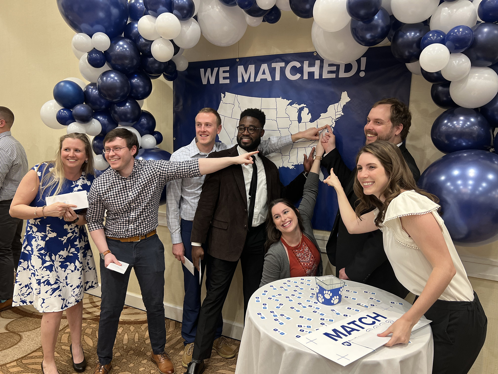 Matching MD students point toward a poster that says We Matched! with a U.S. map on it, with an arch of blue and white balloons above them. 