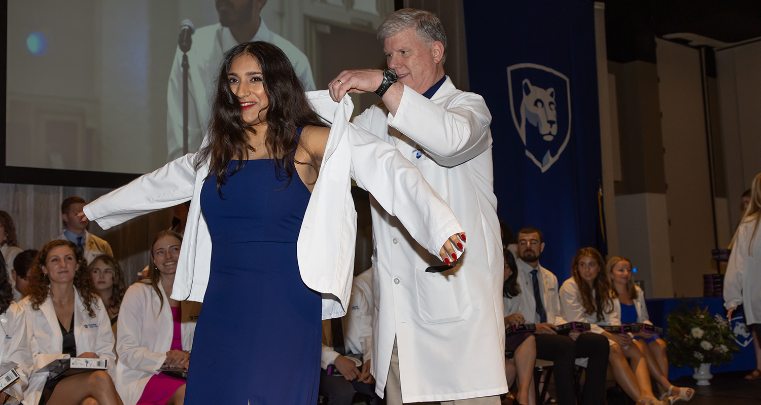 Dr. Michael Flanagan holds up a white coat while an incoming medical student puts it on during the 2024 white coat ceremony