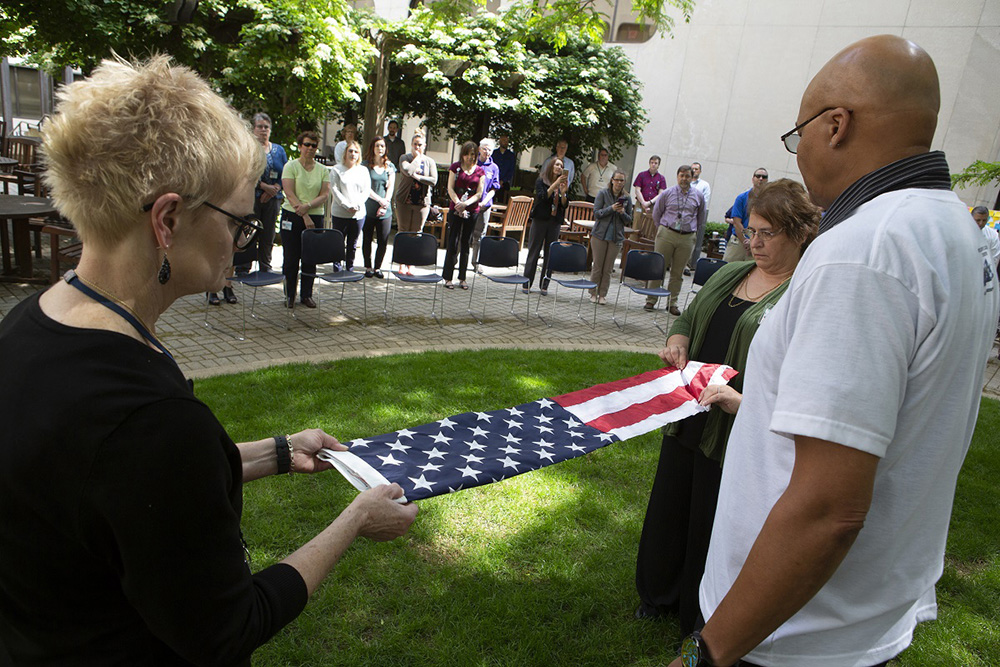 A photo of two people folding an American flag during an outdoors memorial day ceremony, as a group of people watch.