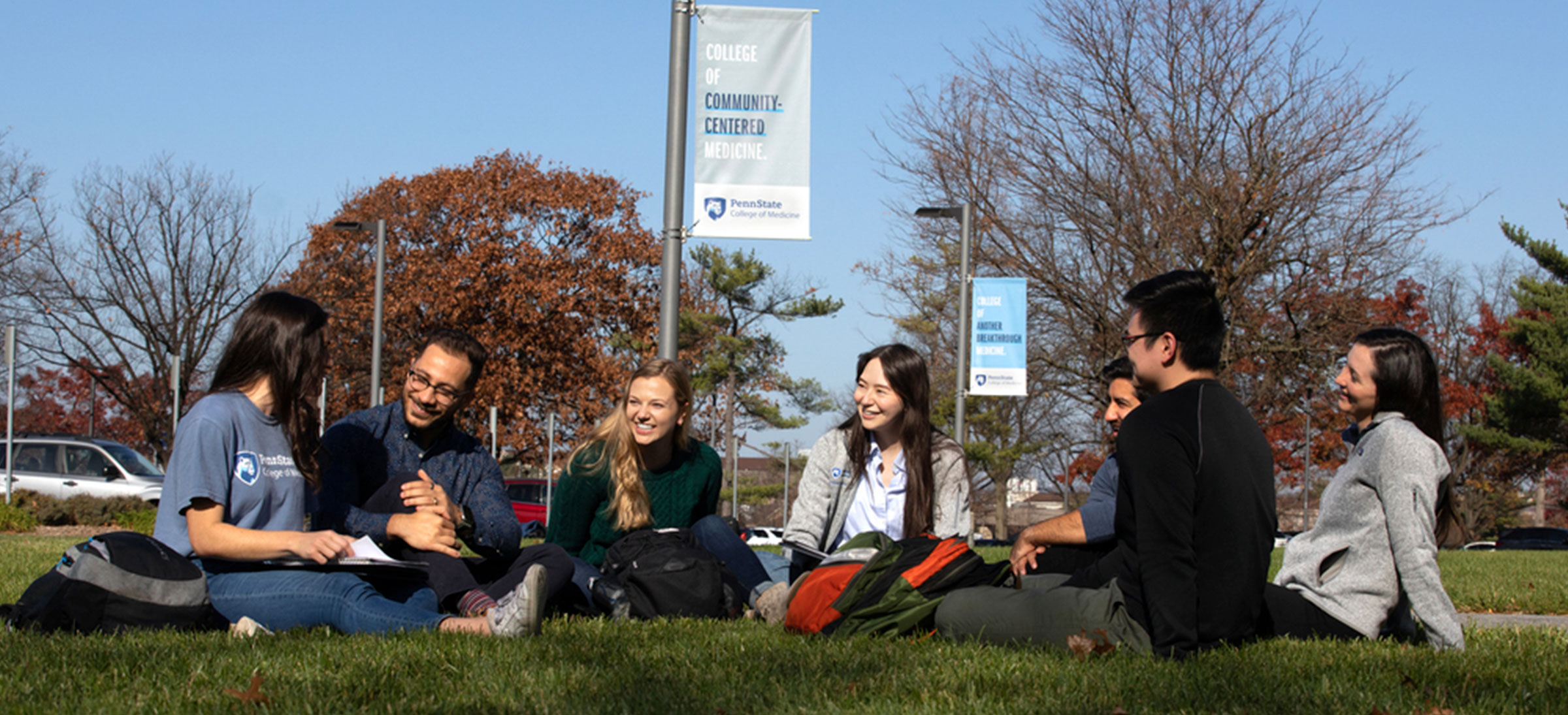 Three Penn State College of Medicine students are seen sitting in an outdoor courtyard in fall 2019, laughing together. A laptop is open on a table in front of them.