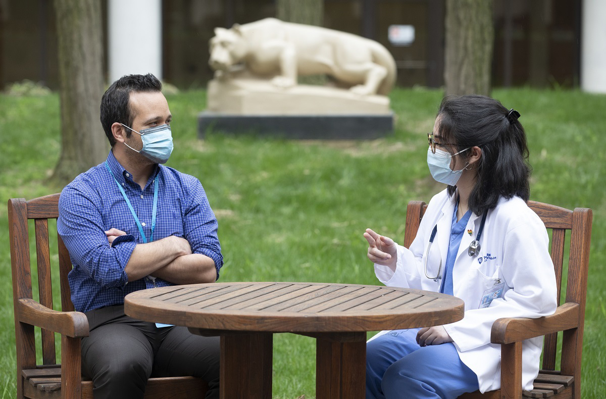Dr. Jonathan Nunez, an internal medicine and infectious disease physician, and Stephanie Hawkins, a second-year medical student, sit outside at a wooden table with a statue of the Penn State Nittany Lion in the background. They both wear face masks and Hawkins is wearing glasses and hospital scrubs under a lab coat.