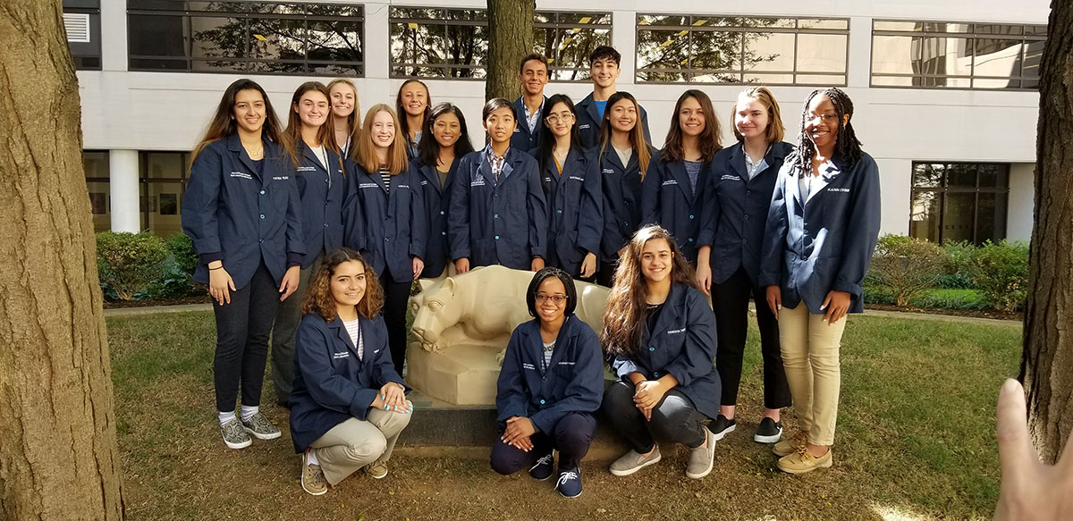 Photo of a group of high school students in blue HCEP coats, surrounding the Nittany Lion statue in the courtyard at the Penn State College of Medicine.
