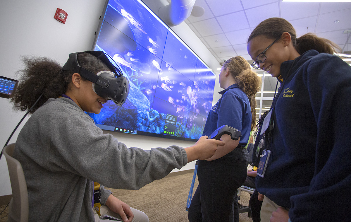 Three students are in a brightly lit classroom. One student is standing, looking at a large video screen which displays an underwater image. The other two students are interacting with each other; one is sitting in a chair, using a virtual reality headset and the other is standing, observing and smiling.