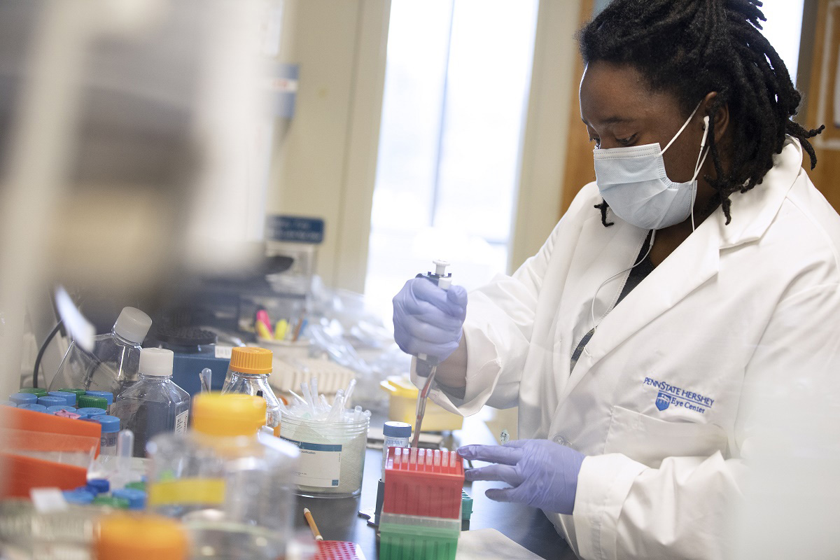 A photo of a PhD/MD student at Penn State College of Medicine who is wearing a White Coat and a PPE, working in a lab.