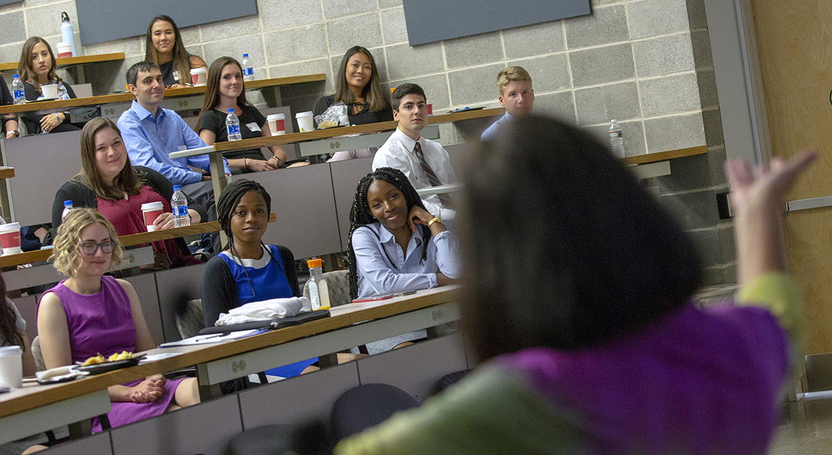 Pictured are medical students of mixed gender and diverse backgrounds who are seated in a portion of a large lecture room and smiling, with a person speaking to them.