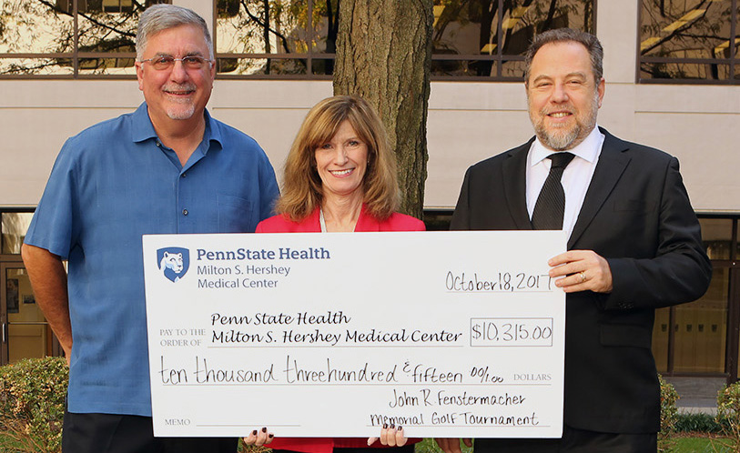 Three people stand in a line, holding a large check made out to Penn State Health Milton S. Hershey Medical Center.