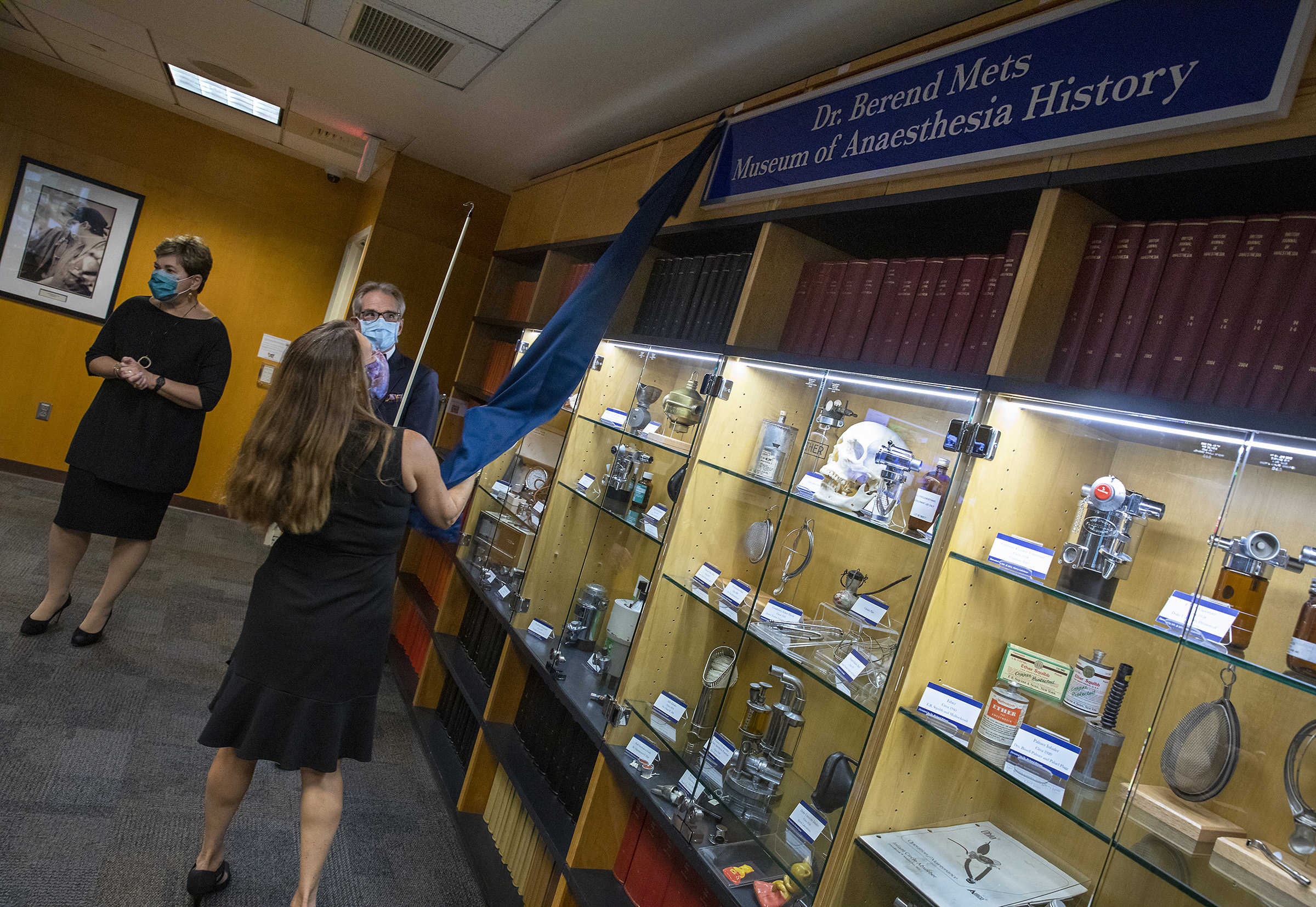 A woman pulls a covering off the sign reading Dr. Berend Mets Museum of Anaesthesia History hanging above a large display case, while a man looks on and a woman stands nearby.