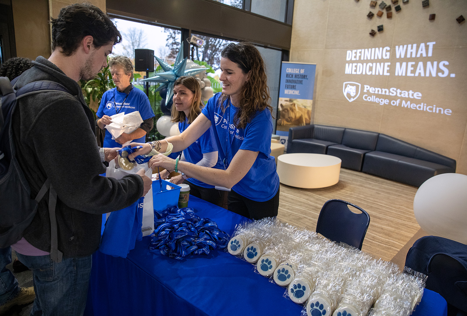 Three people wearing Penn State College of Medicine shirts stand behind a table, handing out items to two people on the other side of the table; there are balloons in the background and Defining What Medicine Means projected on a wall.