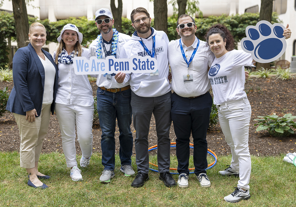 Six people, all wearing white shirts and some wearing white pants and hats, pose holding We are Penn State and paw print signs in the College of Medicine courtyard.
