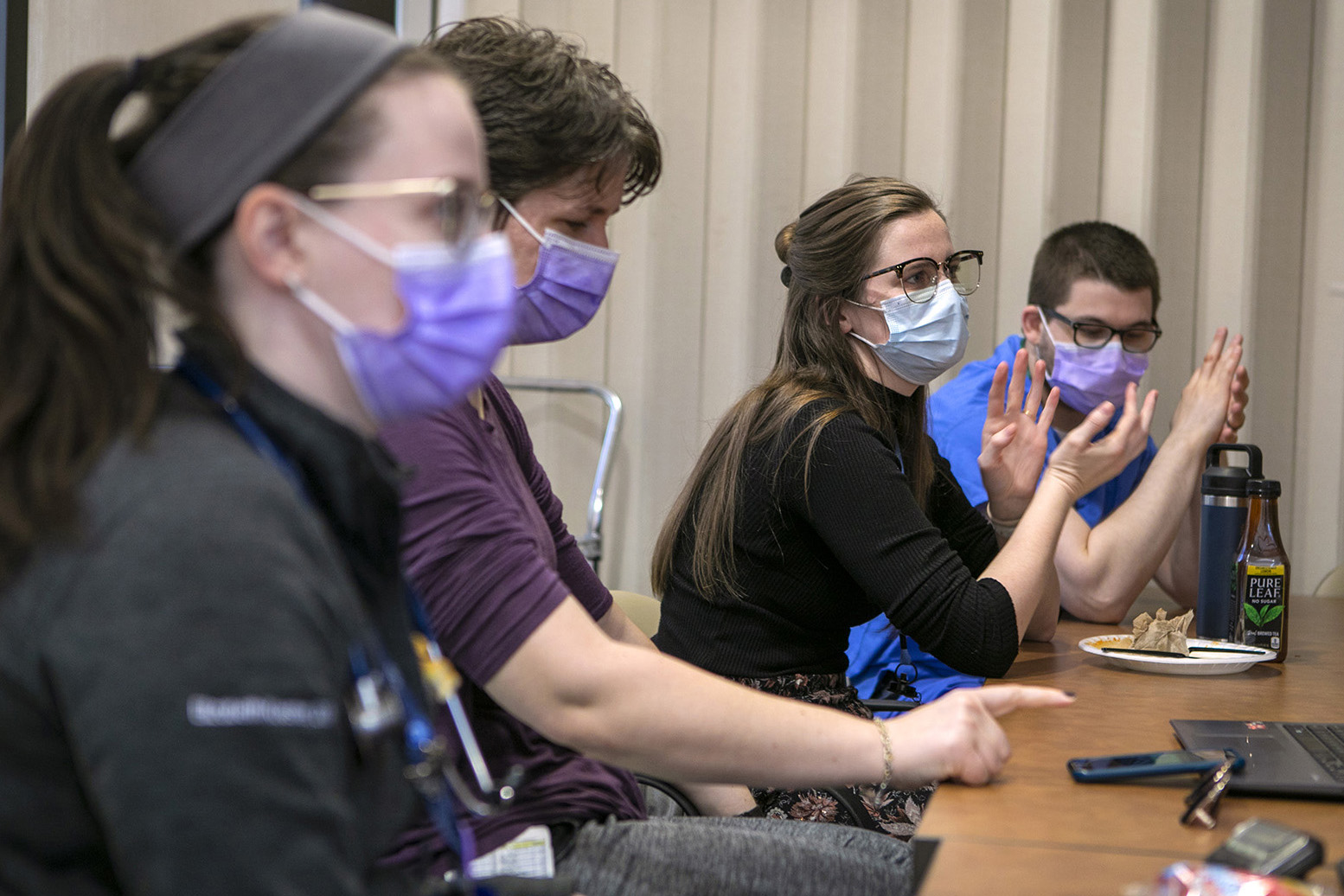 Four residents wearing face masks sit at a table, with the third from the left in focus; she is gesturing and looking ahead.