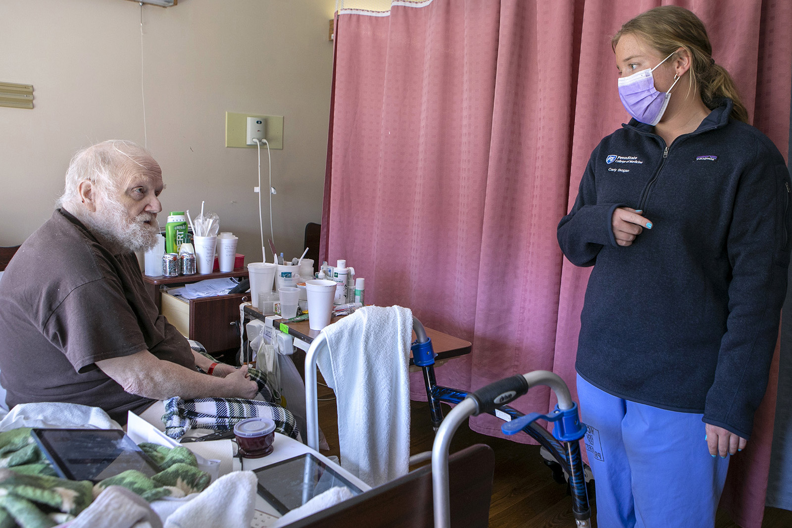  First-year medical student Carly Brogan, standing at right, talks with patient Robert Gibbs, who is sitting in a retirement village room. 