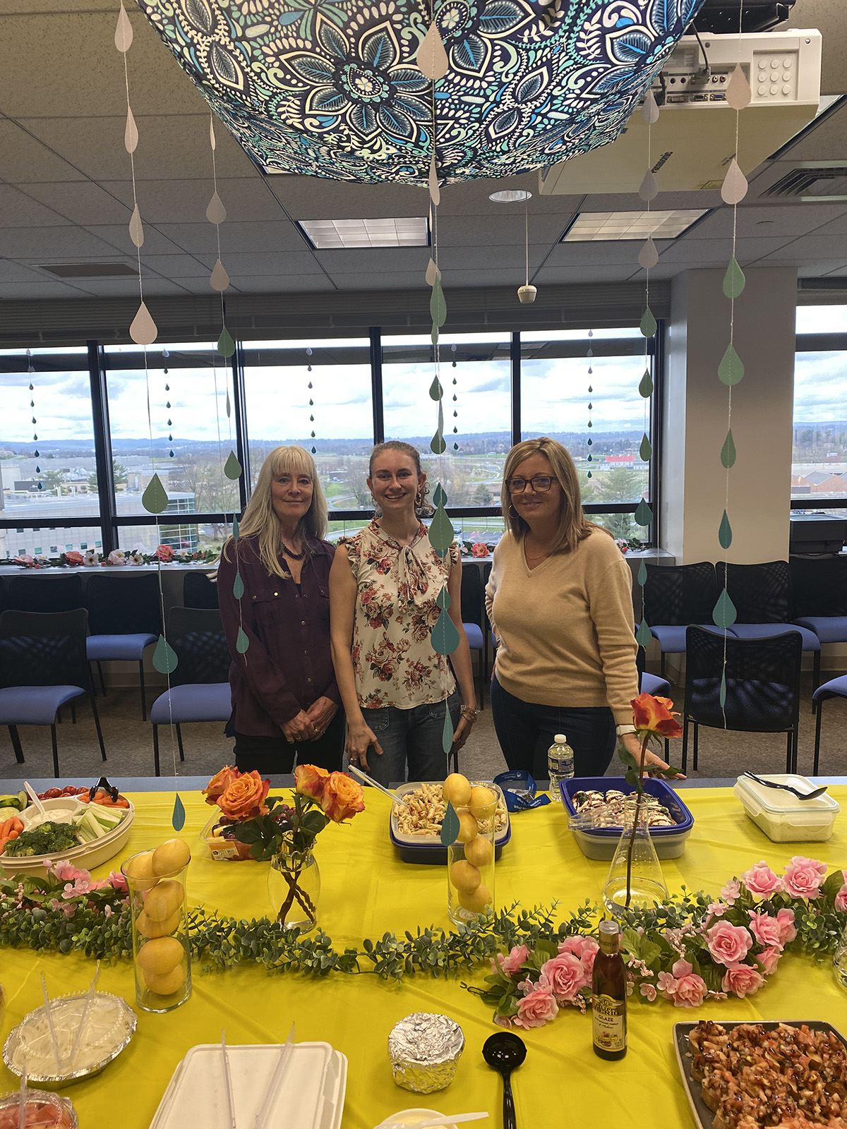Three women stand behind a table with food and decorations with an April Showers Bring May Flowers theme.